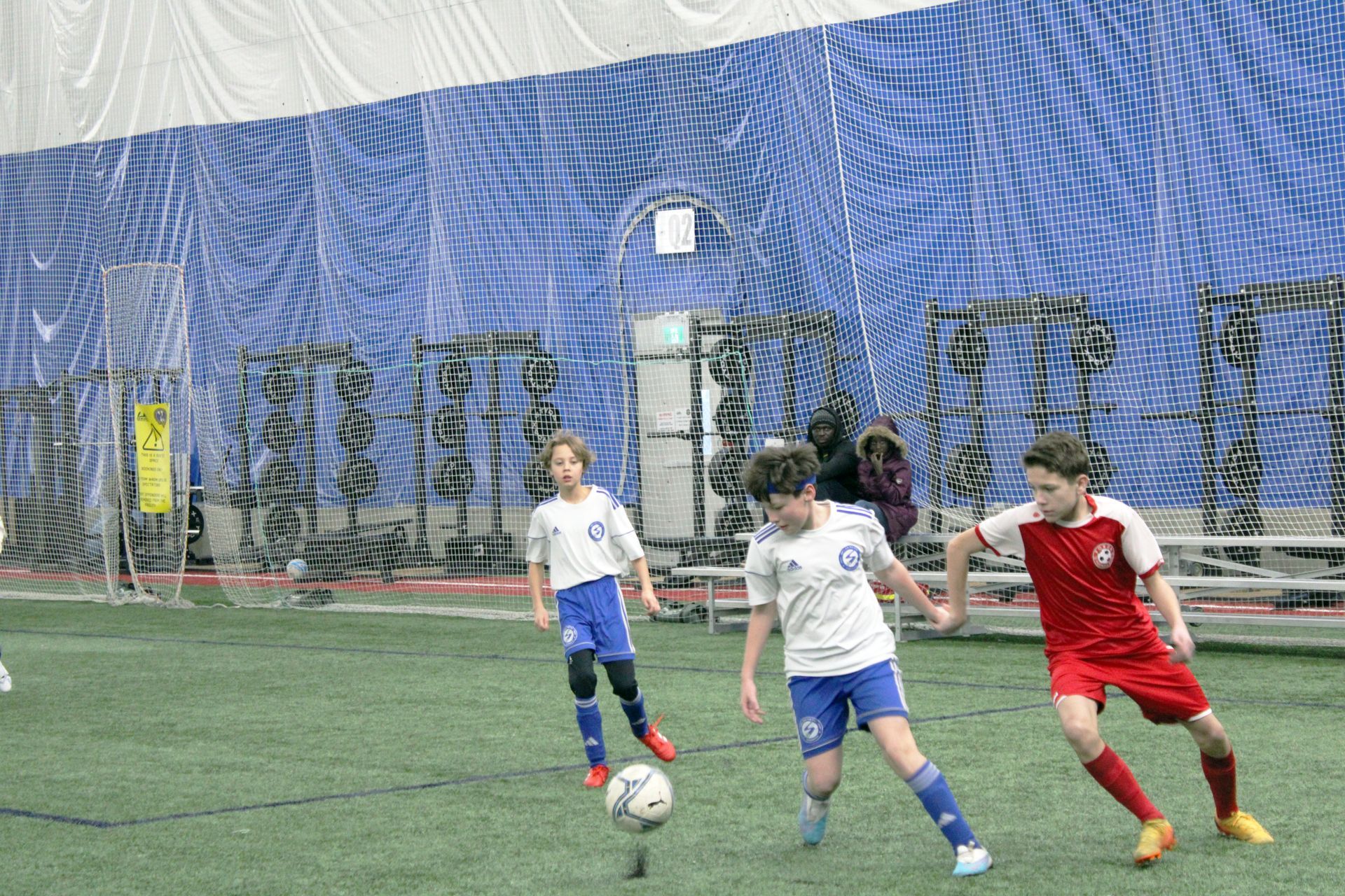 Three children in soccer uniforms compete for a ball on an indoor turf field with a blue net backdrop.