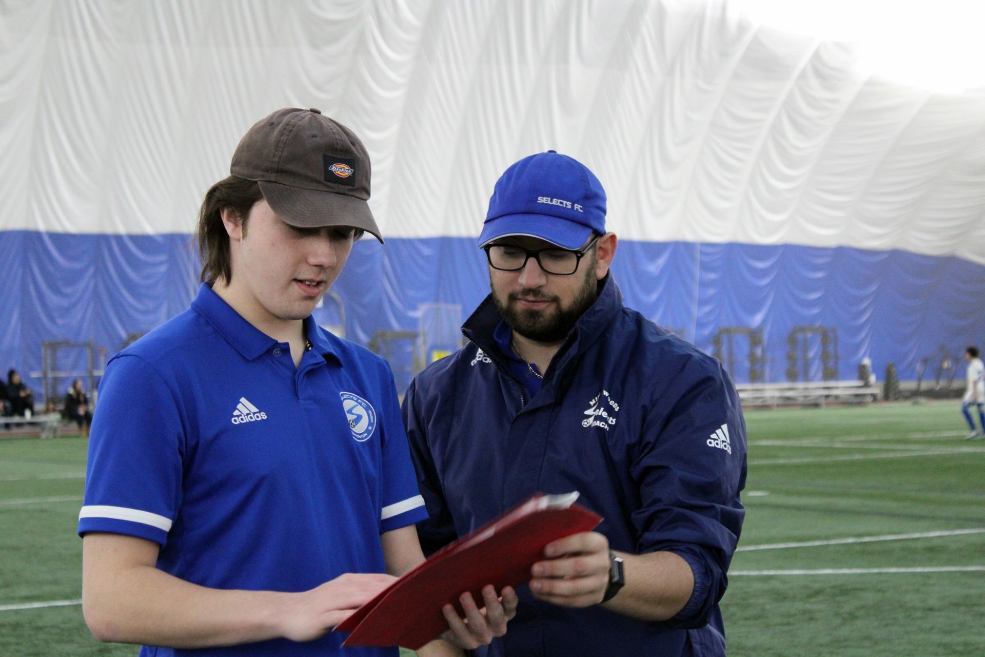 Two people in blue athletic gear stand on an indoor turf field, reviewing notes on a red clipboard.