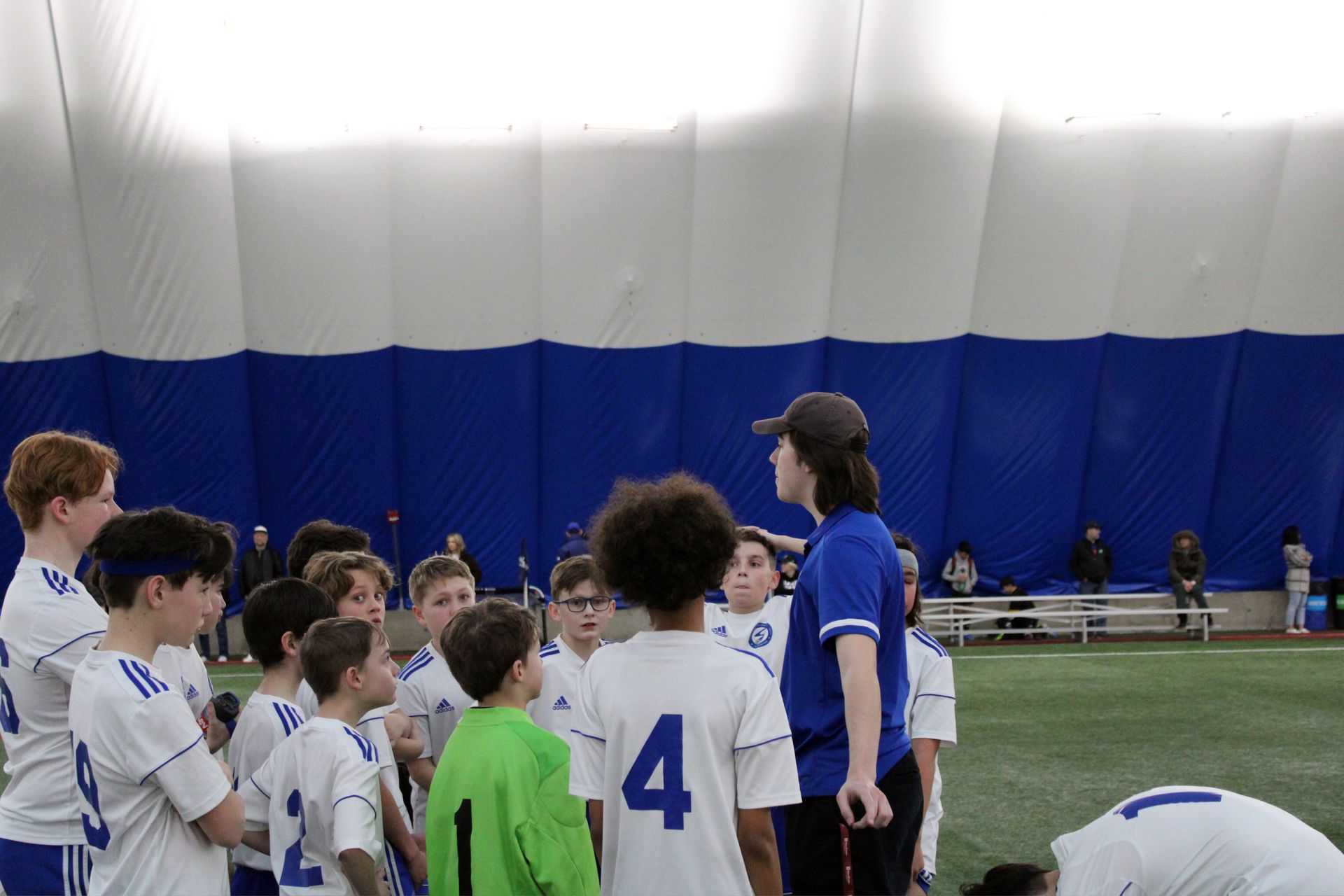 A coach stands in a huddle with a team of soccer players wearing white jerseys on an indoor field.