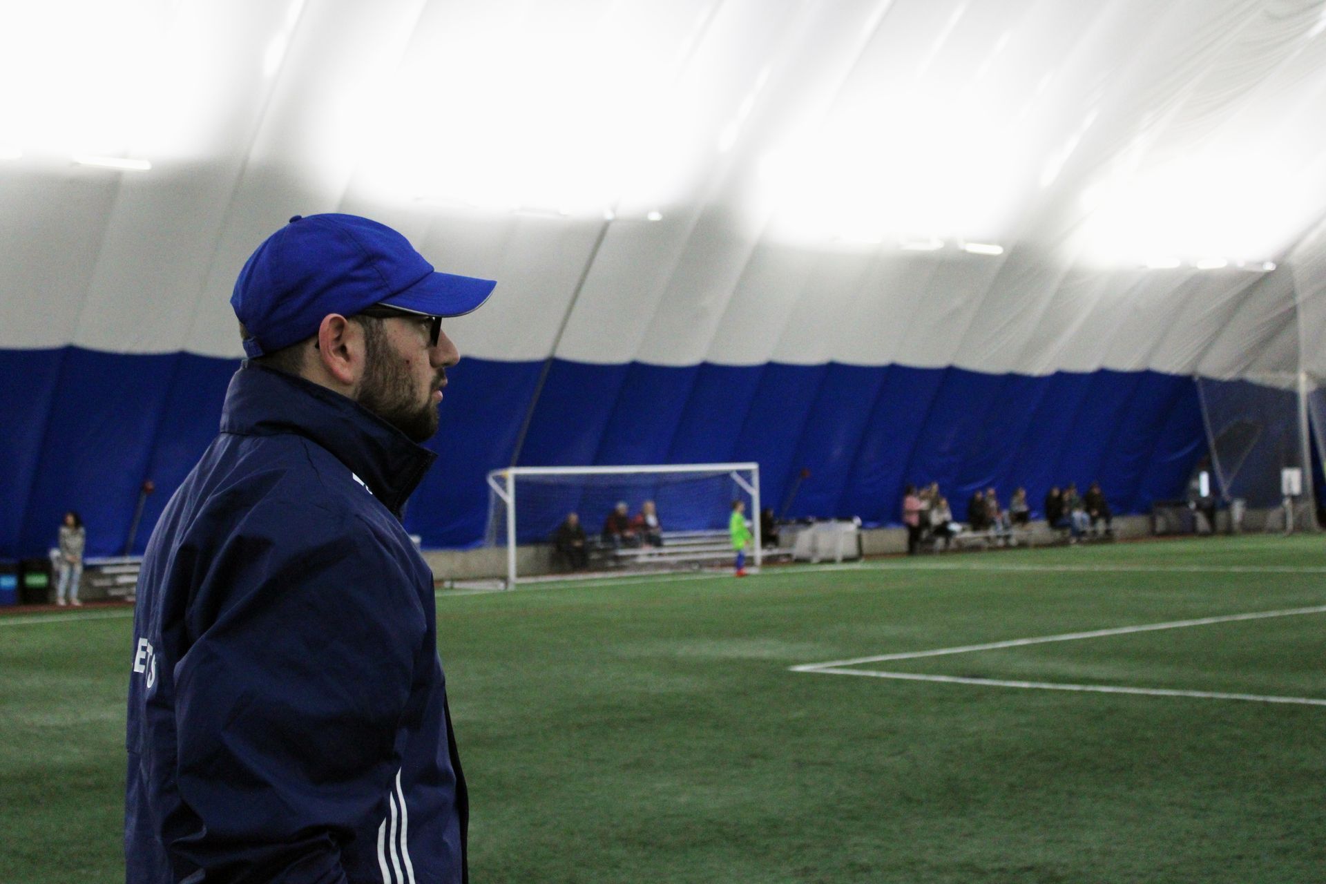 A person wearing a blue cap and jacket stands on an indoor soccer field, watching players by the goal.