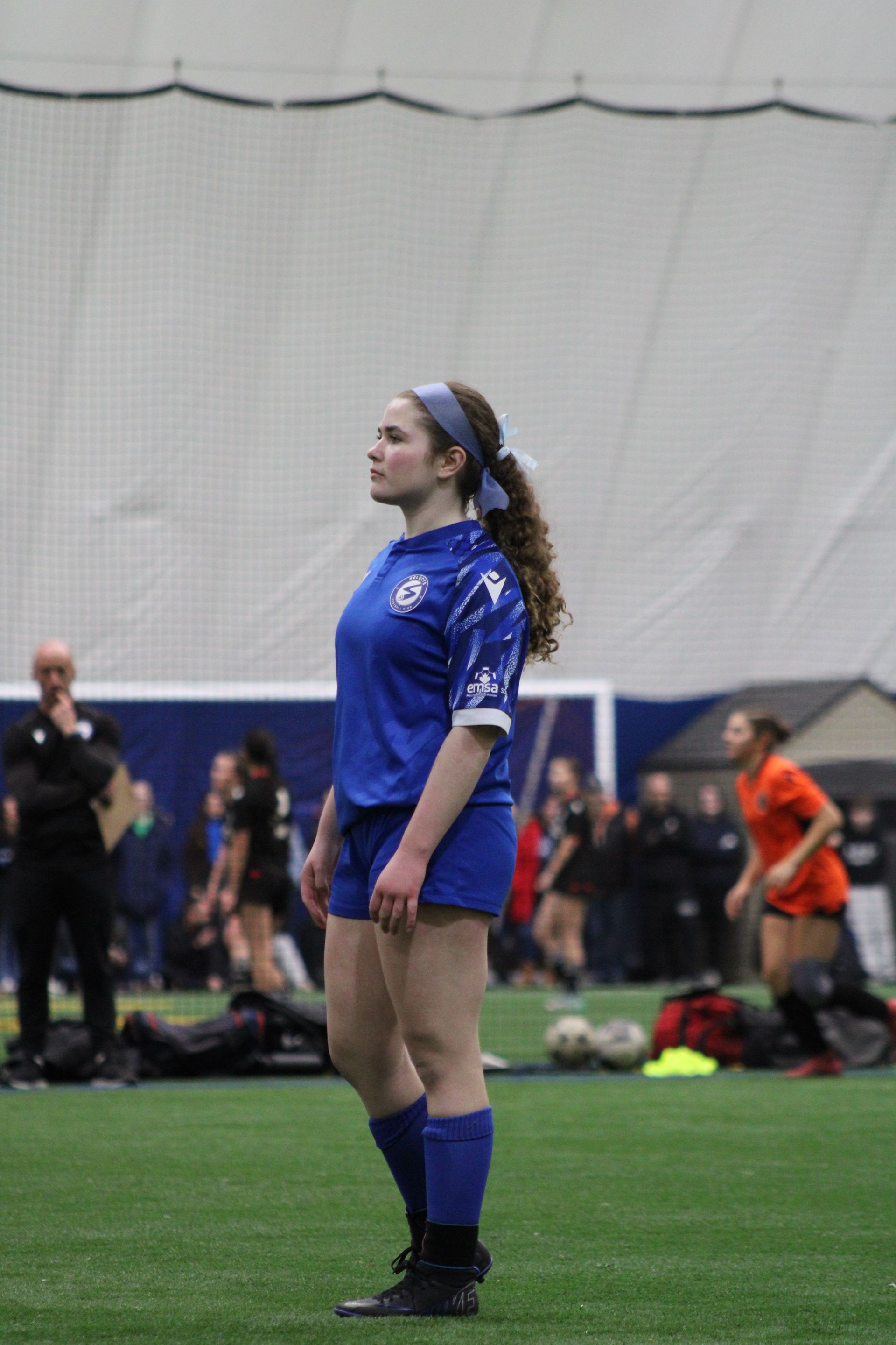 A player in a blue soccer uniform stands on an indoor field, looking to the side with teammates in the background.