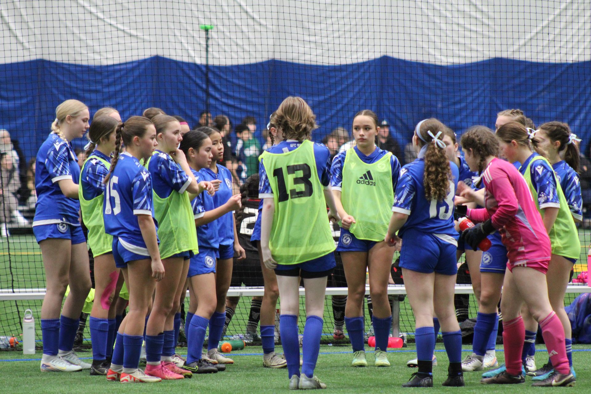 A youth soccer team in blue uniforms stands in a huddle on a green indoor field, some wearing bright yellow practice vests.