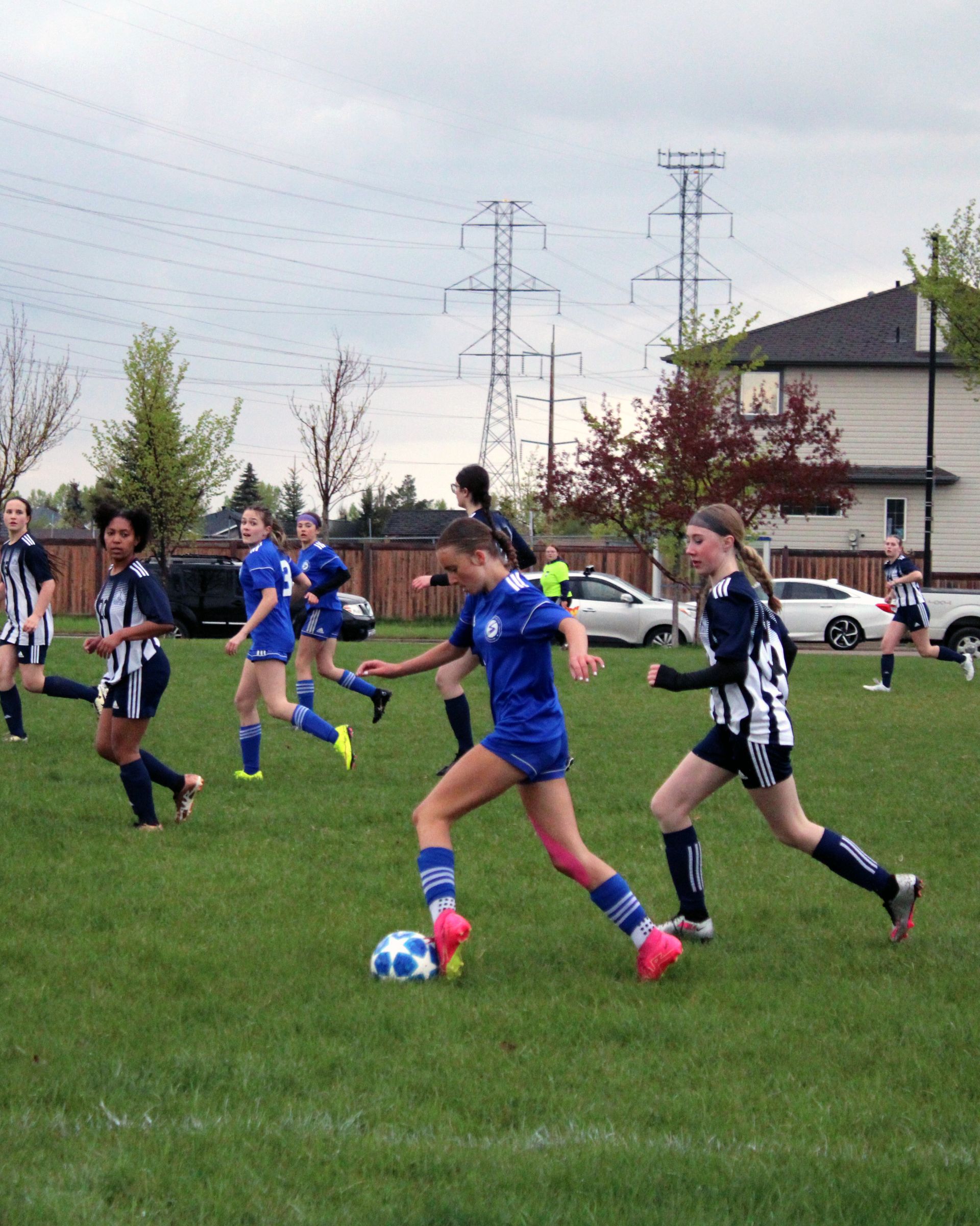 Players in blue and striped uniforms play a soccer game on a grass field under a power line near a residential area.