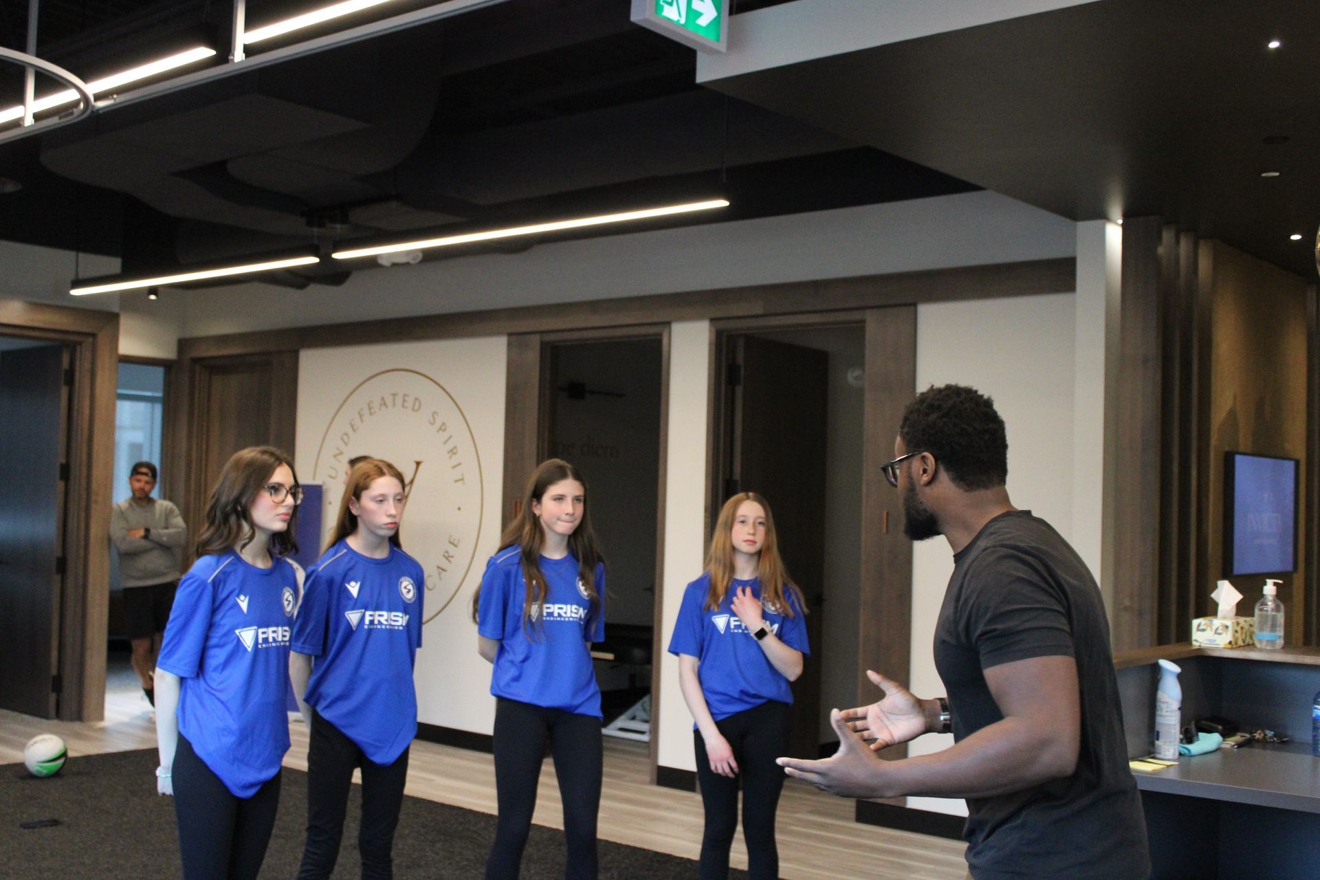 A man gestures while speaking to four people wearing blue jerseys in a modern, indoor office-like space.
