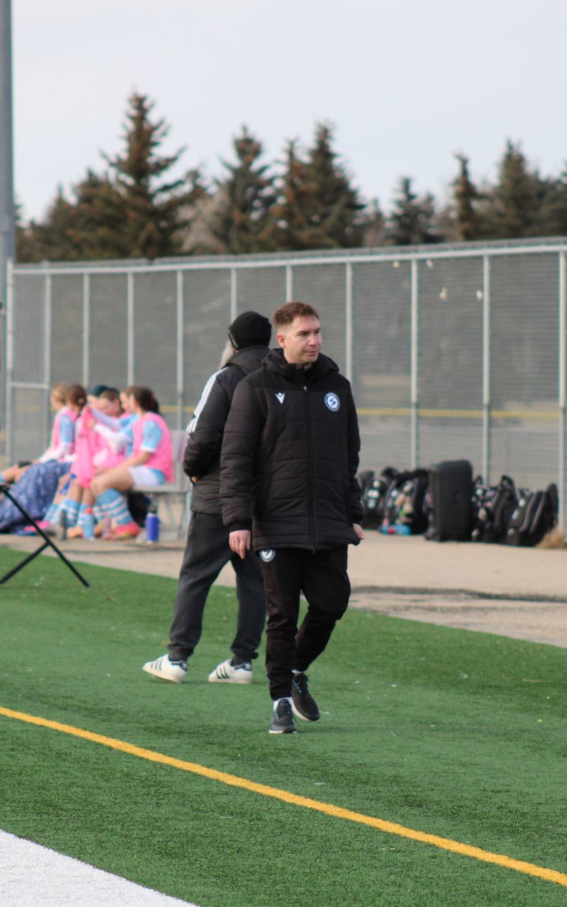A coach with arms crossed, wearing a black shirt with a blue emblem, stands in front of a soccer goal net.