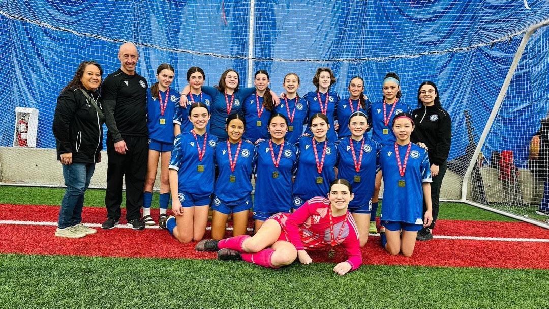 A soccer team in blue uniforms stands with two coaches on a field, one player reclining in front in a red uniform.