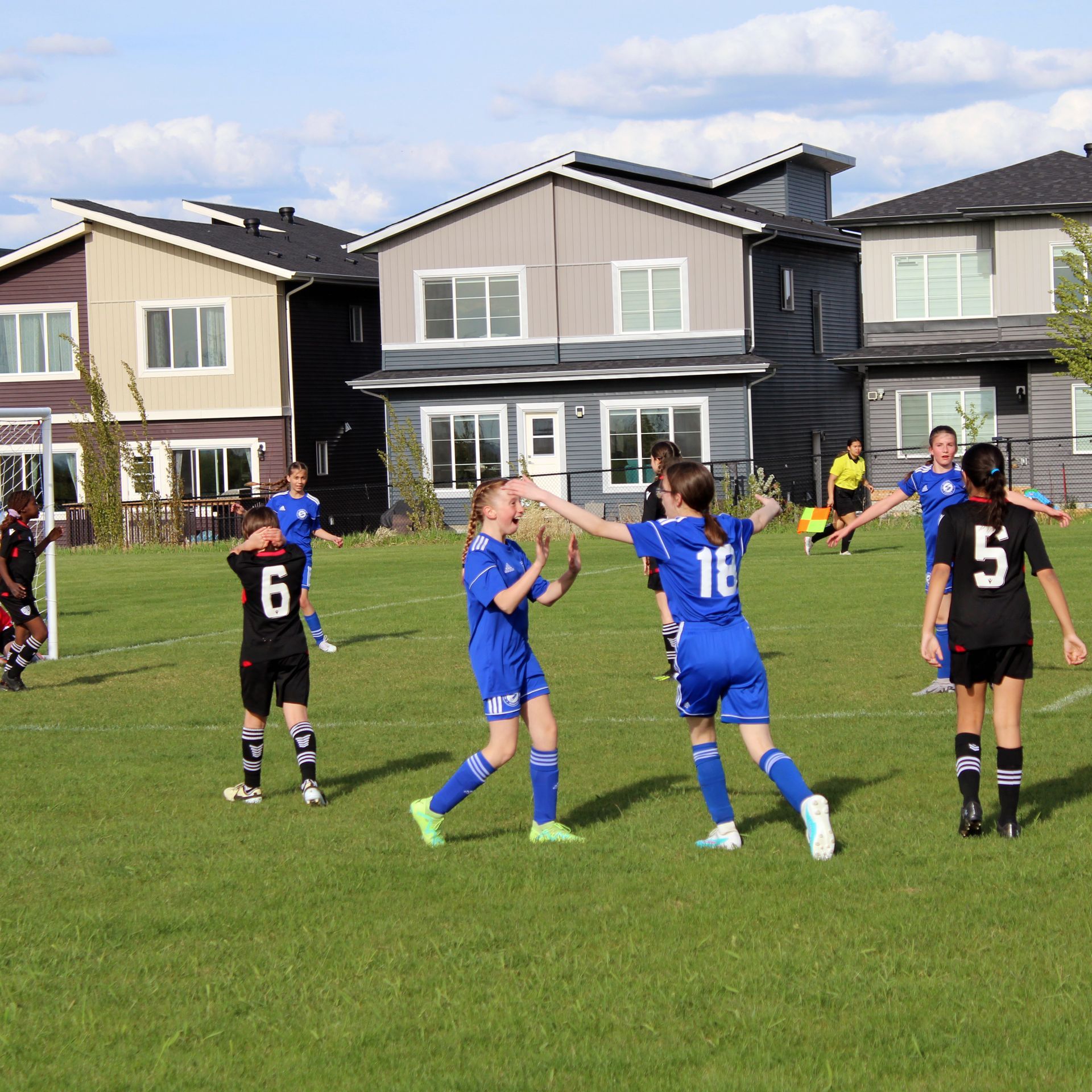 Soccer players in blue and black uniforms celebrate a goal on a grass field in front of residential houses.