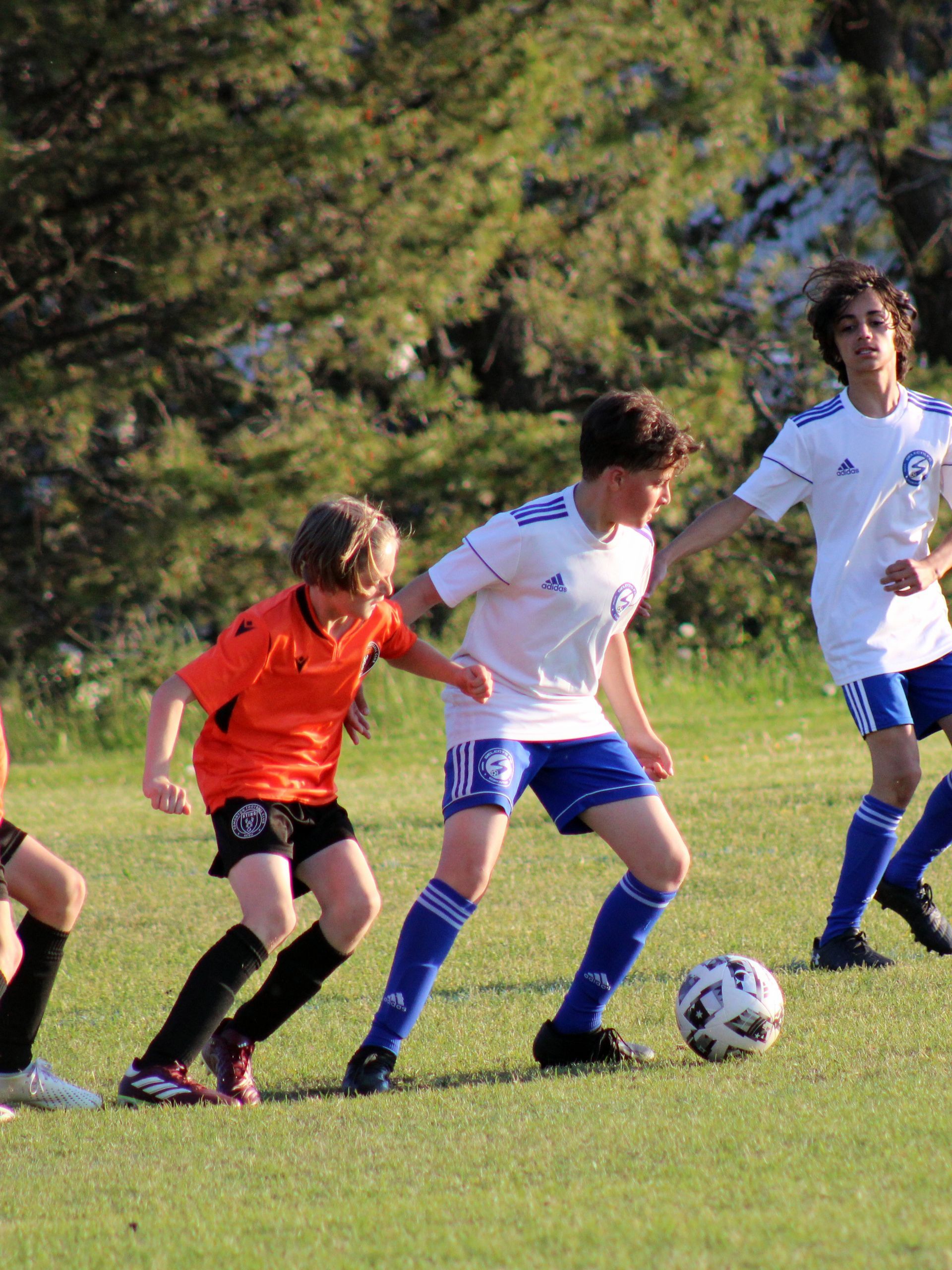 A player in an orange jersey dribbles a soccer ball while being closely defended by two players in white and blue uniforms.