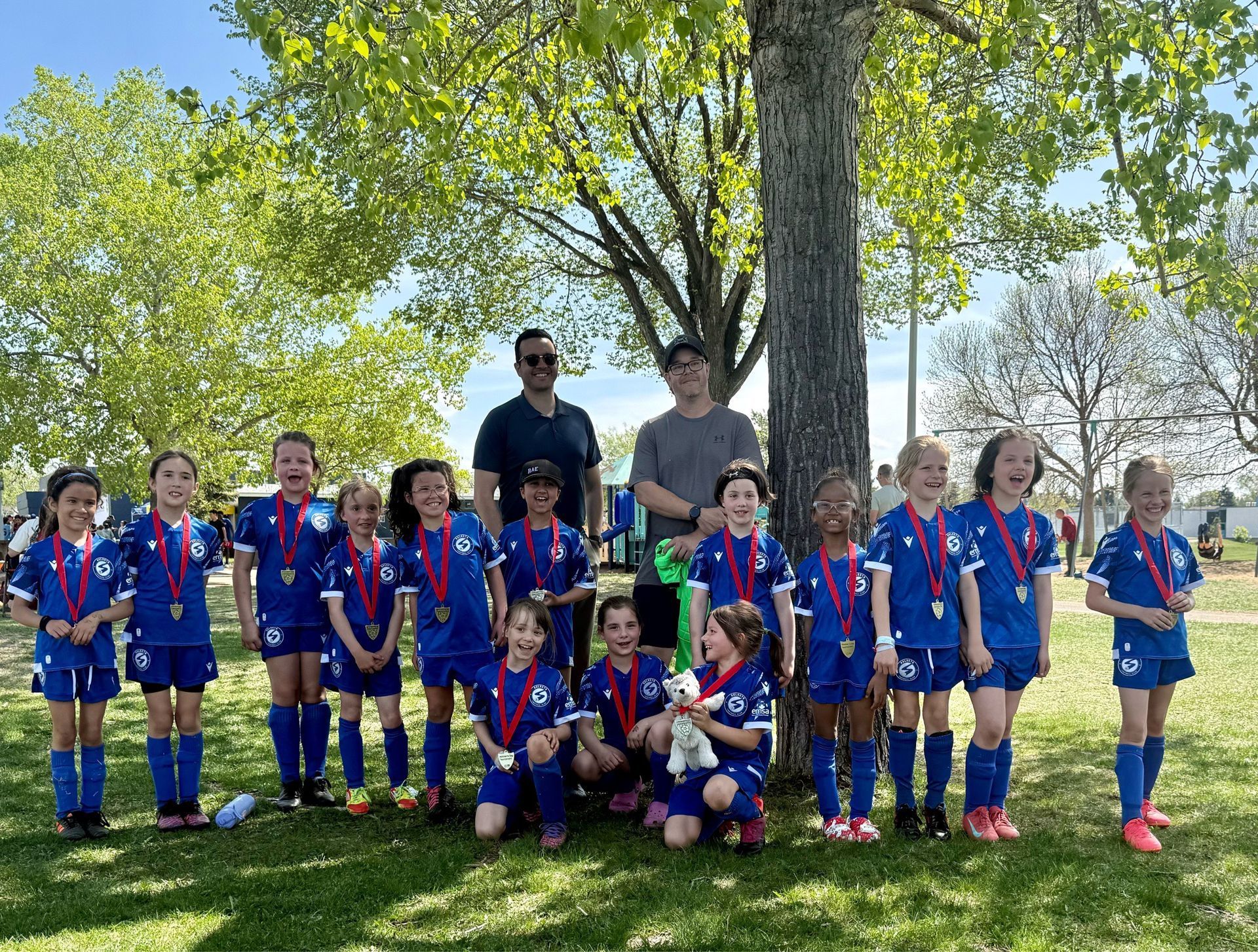 A youth soccer team in blue uniforms stands with two coaches under a large tree in a park on a sunny day.