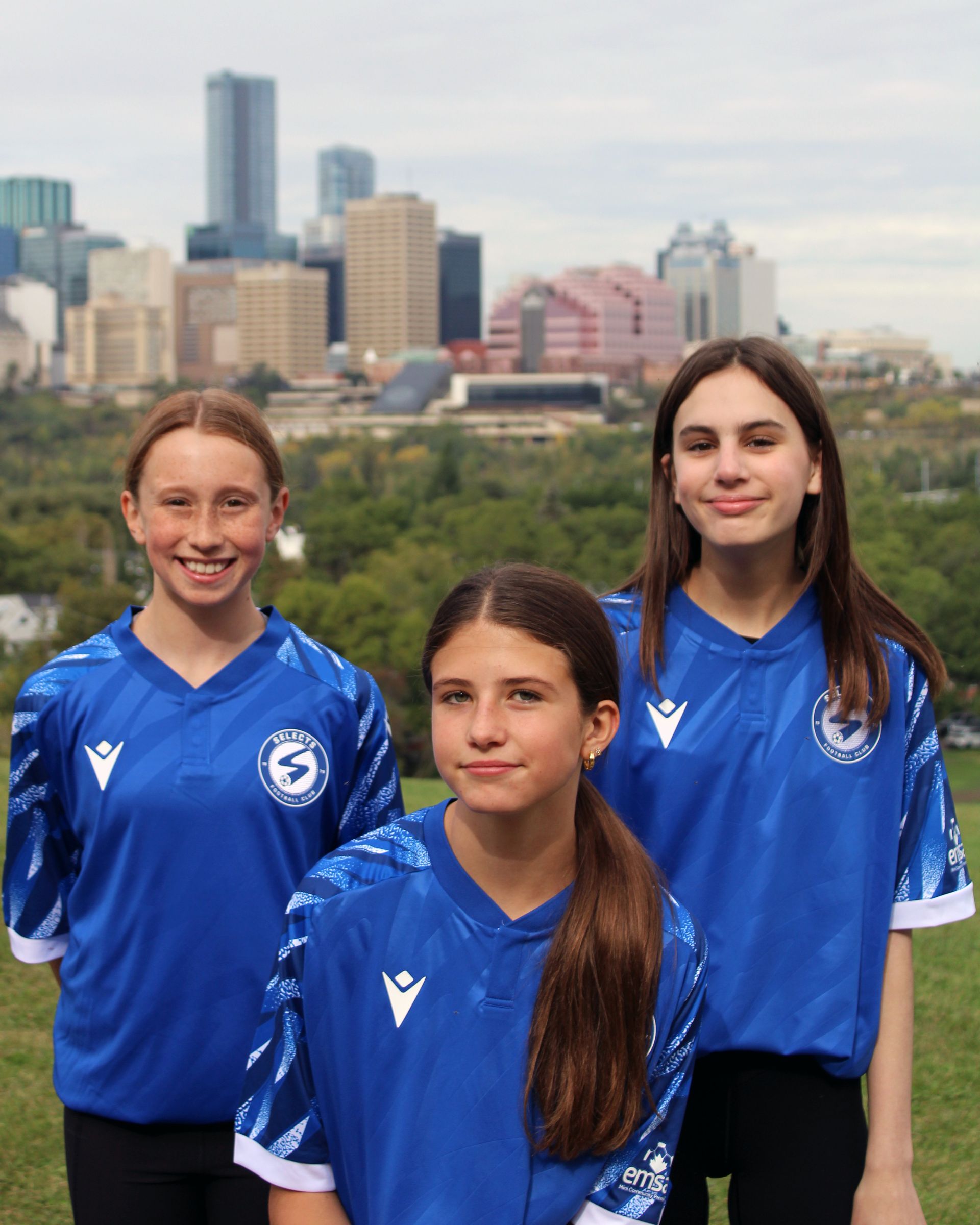 Three people in matching blue soccer jerseys stand outdoors with a city skyline in the background.