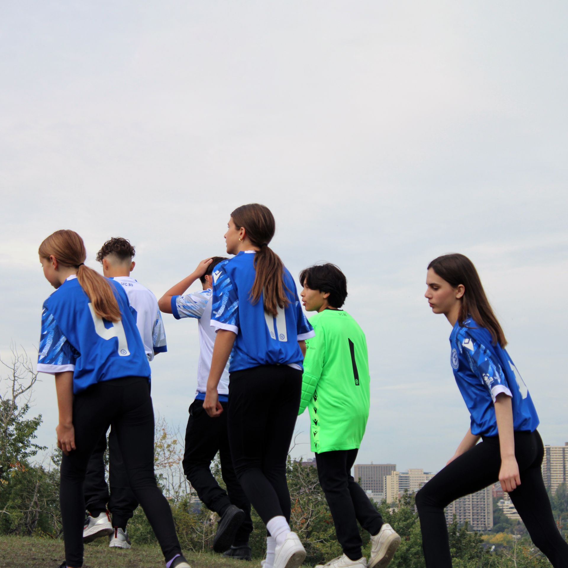 A group of individuals in soccer jerseys and athletic wear walk uphill against a cloudy, bright sky.