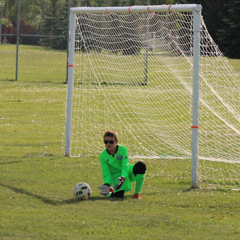 A goalie in a bright green uniform and sunglasses kneeling on a grass field, ready to block a soccer ball in front of a net.