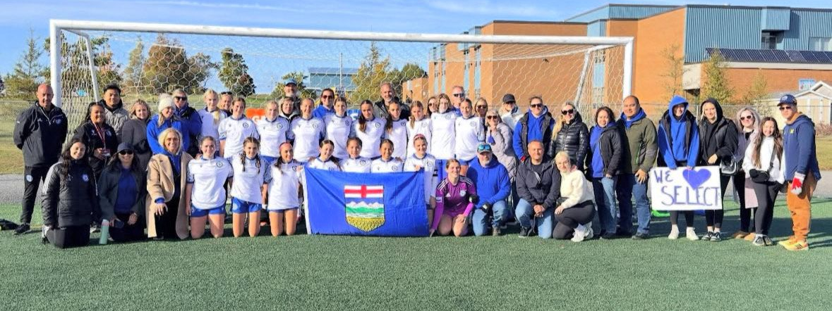 A large soccer team poses with a Saskatchewan provincial flag on a field in front of a goal net and school building.