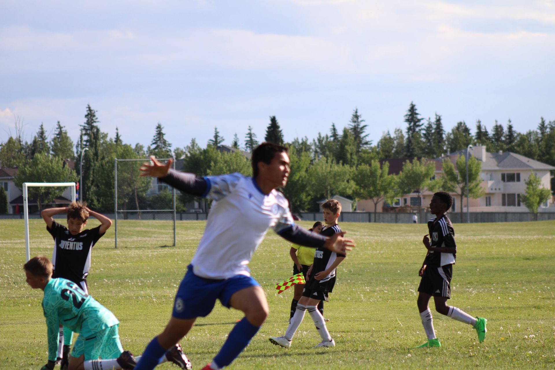 A soccer player in a white jersey celebrates a goal on a field with teammates and an opponent in the background.