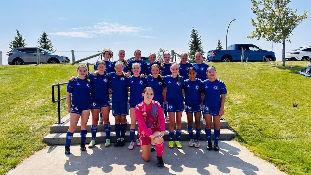A soccer team in blue uniforms poses for a photo on outdoor stairs on a sunny day, with a goalie kneeling in front.