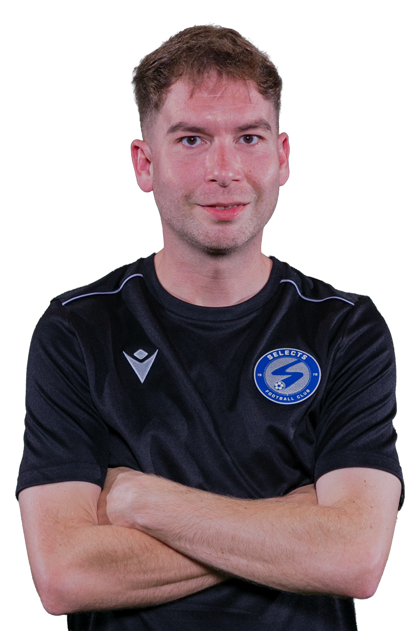 A coach with arms crossed, wearing a black shirt with a blue emblem, stands in front of a soccer goal net.