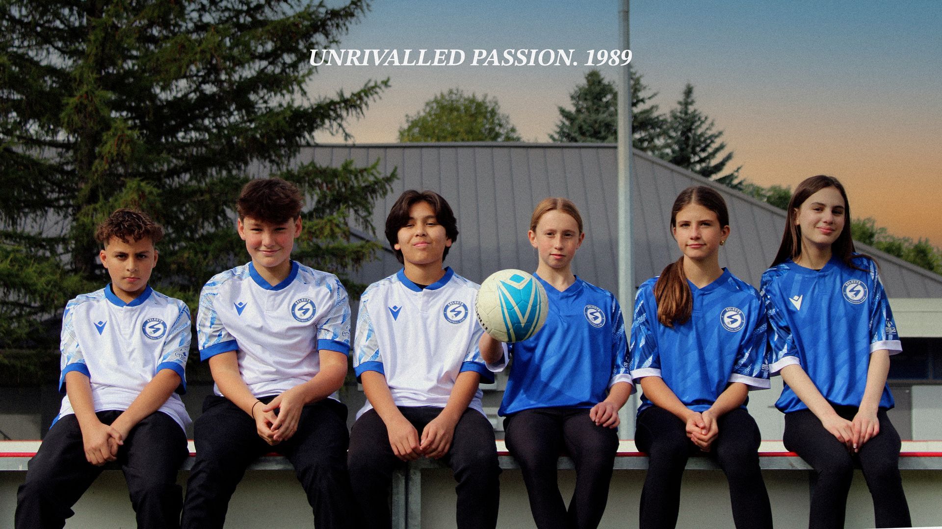 Six youth in athletic jerseys sit side-by-side on a bench in front of a tree, one holding a soccer ball.