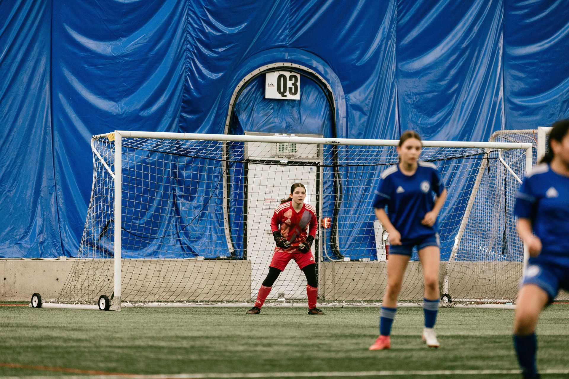A soccer goalkeeper stands in goal wearing a red jersey, with teammates in blue jerseys nearby on an indoor field.