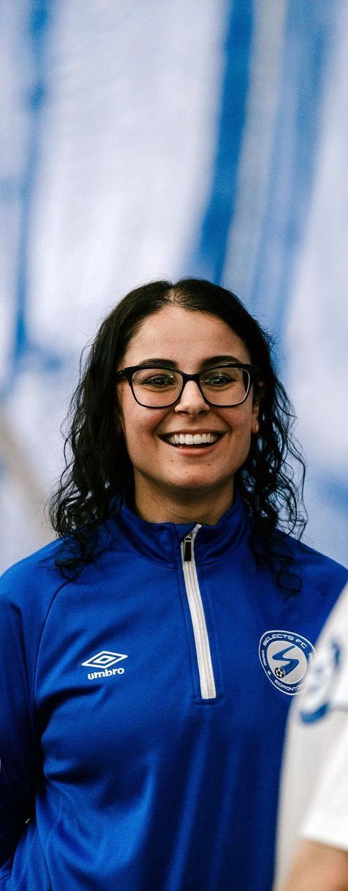A smiling person with dark curly hair and glasses, wearing a blue zip-up athletic jacket with a white logo.