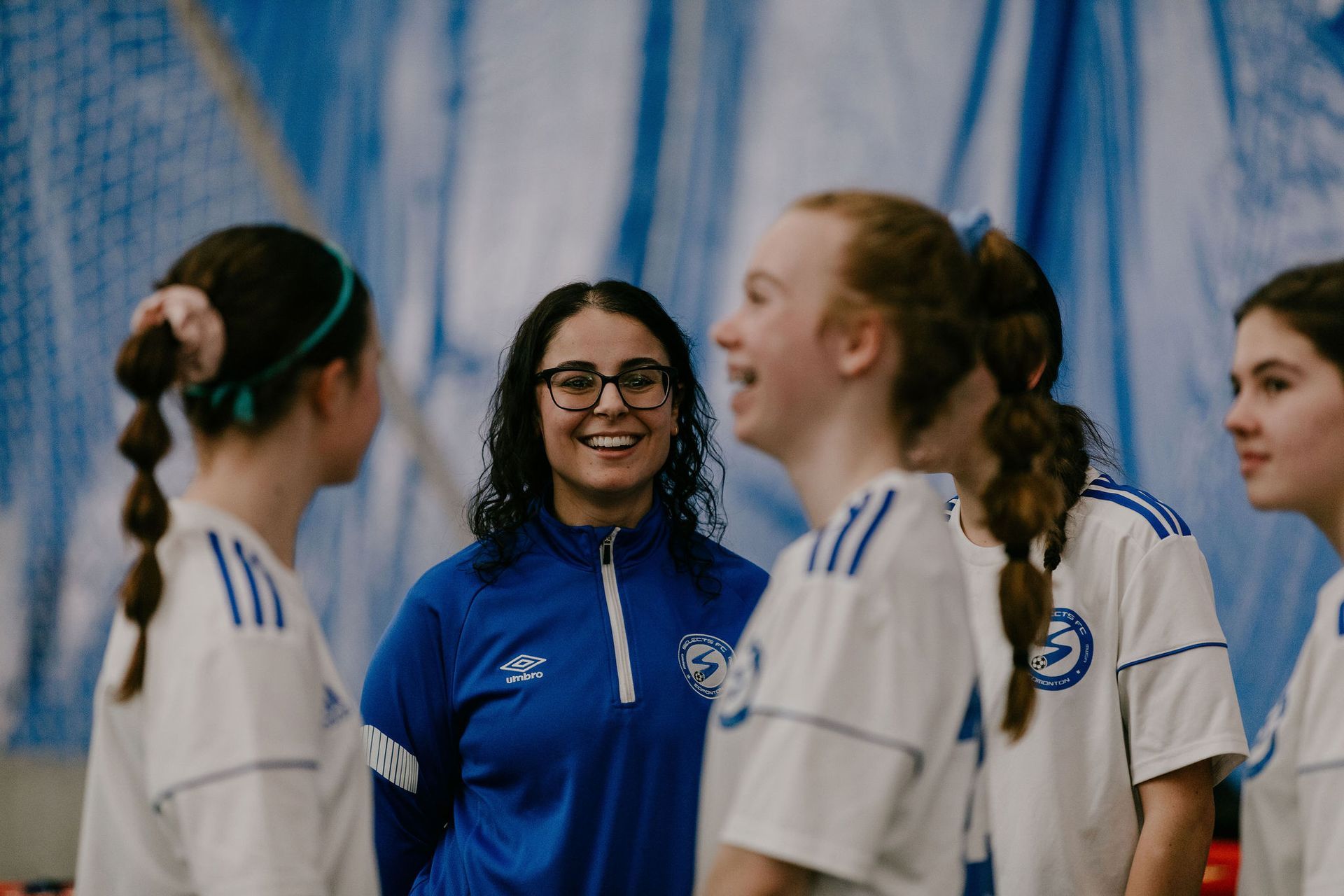 A coach in a blue jacket smiles while talking to a group of athletes in white jerseys at an indoor facility.