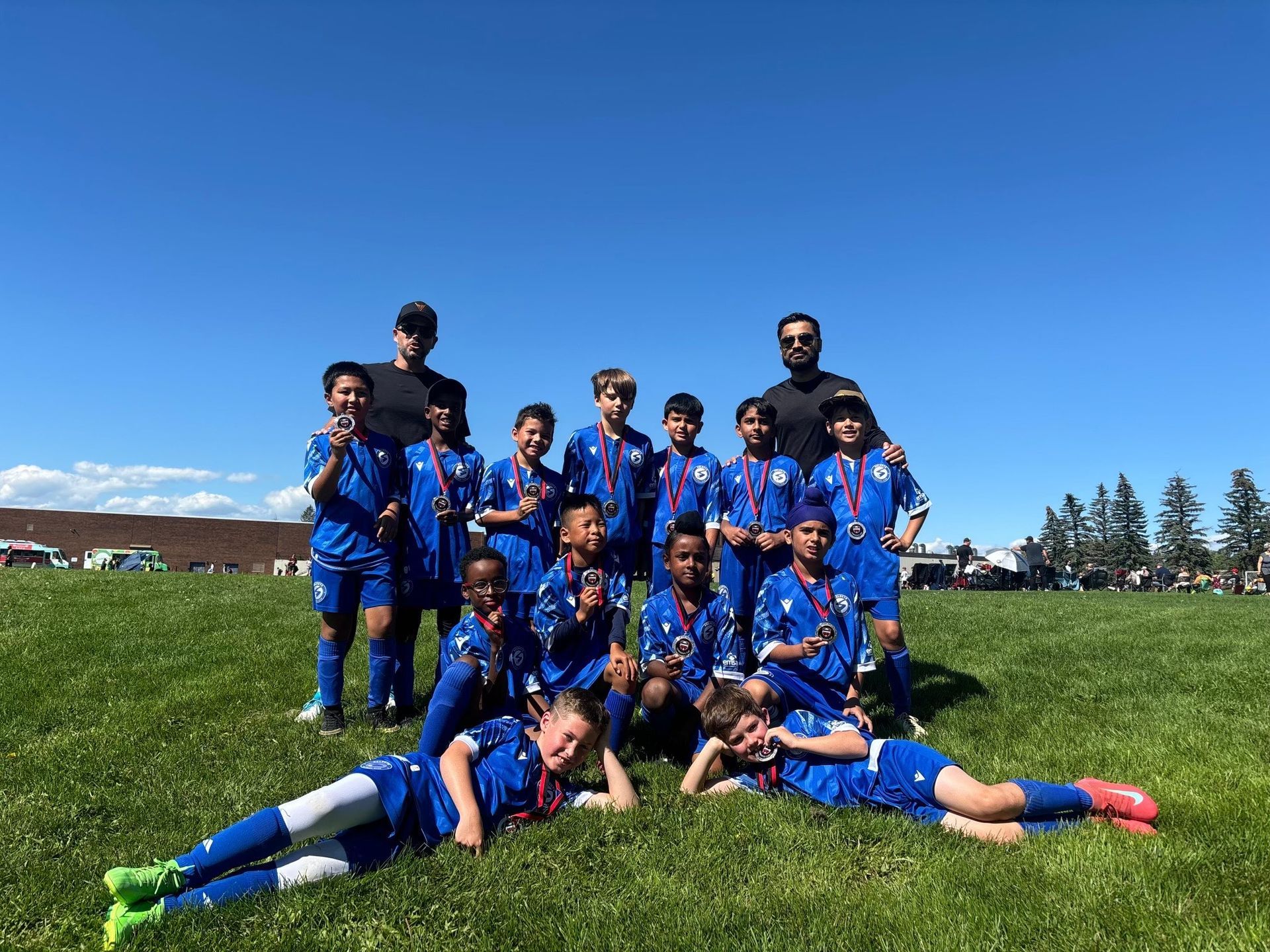 A youth soccer team in blue uniforms and two coaches pose for a photo on a grassy field under a clear blue sky.