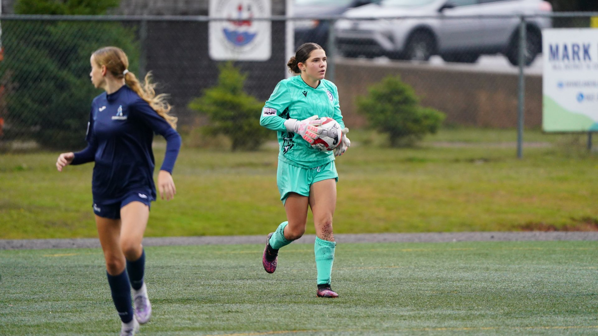 A soccer goalkeeper in a teal kit holds a ball while running on a turf field, as a teammate in dark blue gear runs nearby.