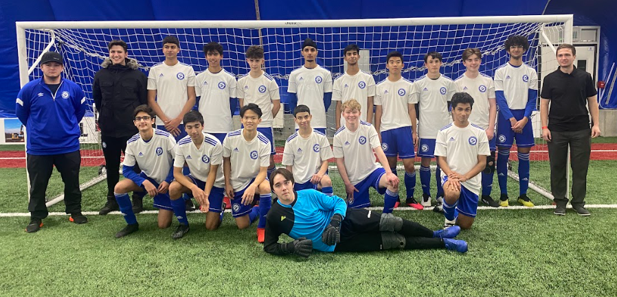 A soccer team in white uniforms and a goalie in blue stand in front of a goal on an indoor turf field with two coaches.