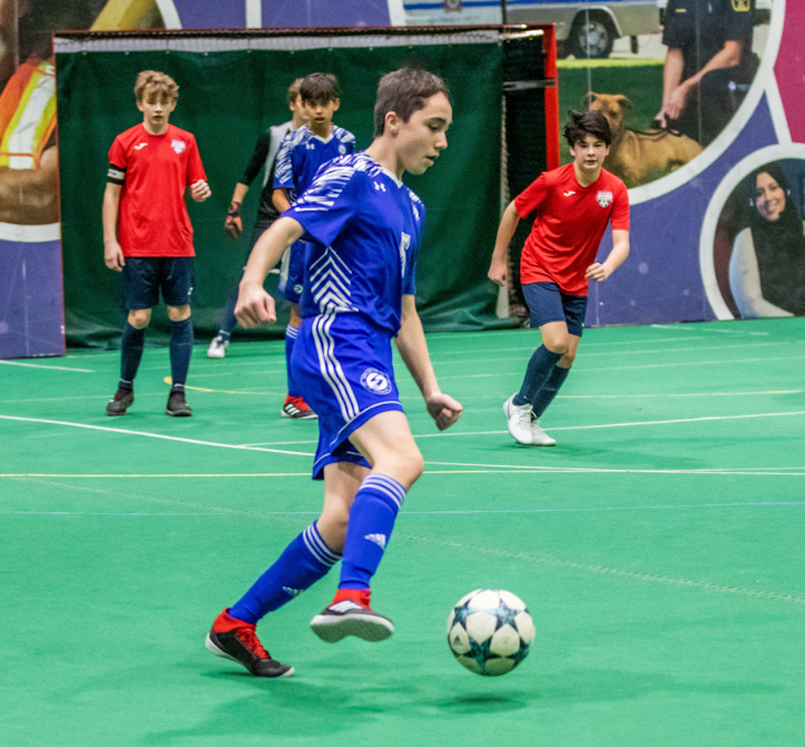 A youth soccer player in a blue uniform dribbles a soccer ball on an indoor field, with opponents in red jerseys nearby.