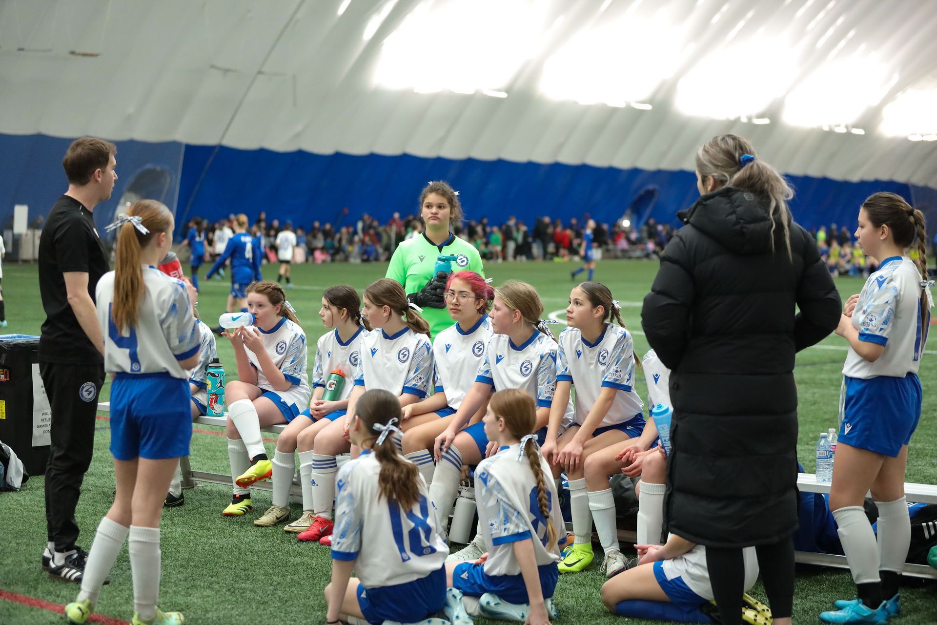 A youth soccer team sits on a bench in an indoor sports facility while coaches speak to them during a game.