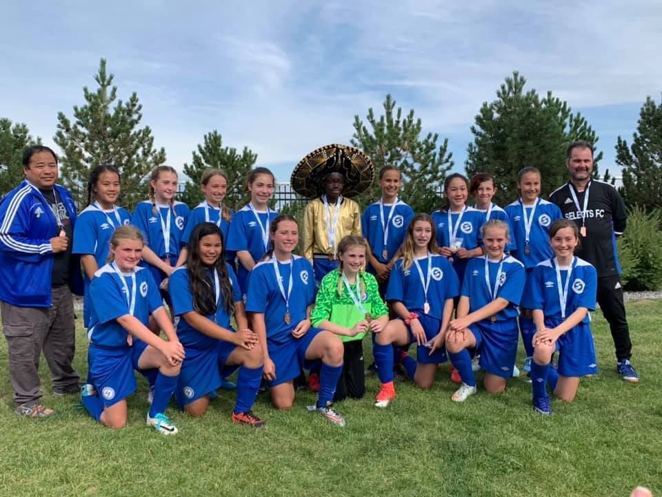 A youth soccer team in blue uniforms stands on a grassy field with two coaches, one wearing a large sombrero.