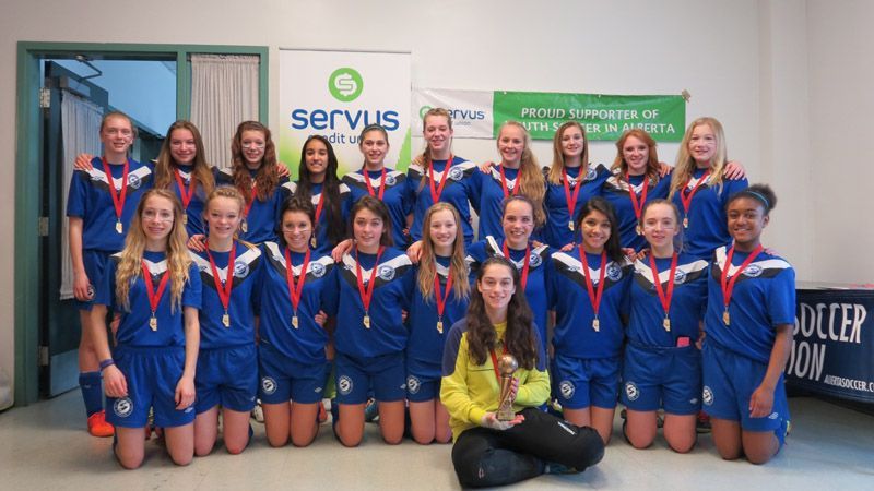 A soccer team in blue uniforms stands together indoors, wearing medals, with one player seated holding a trophy.