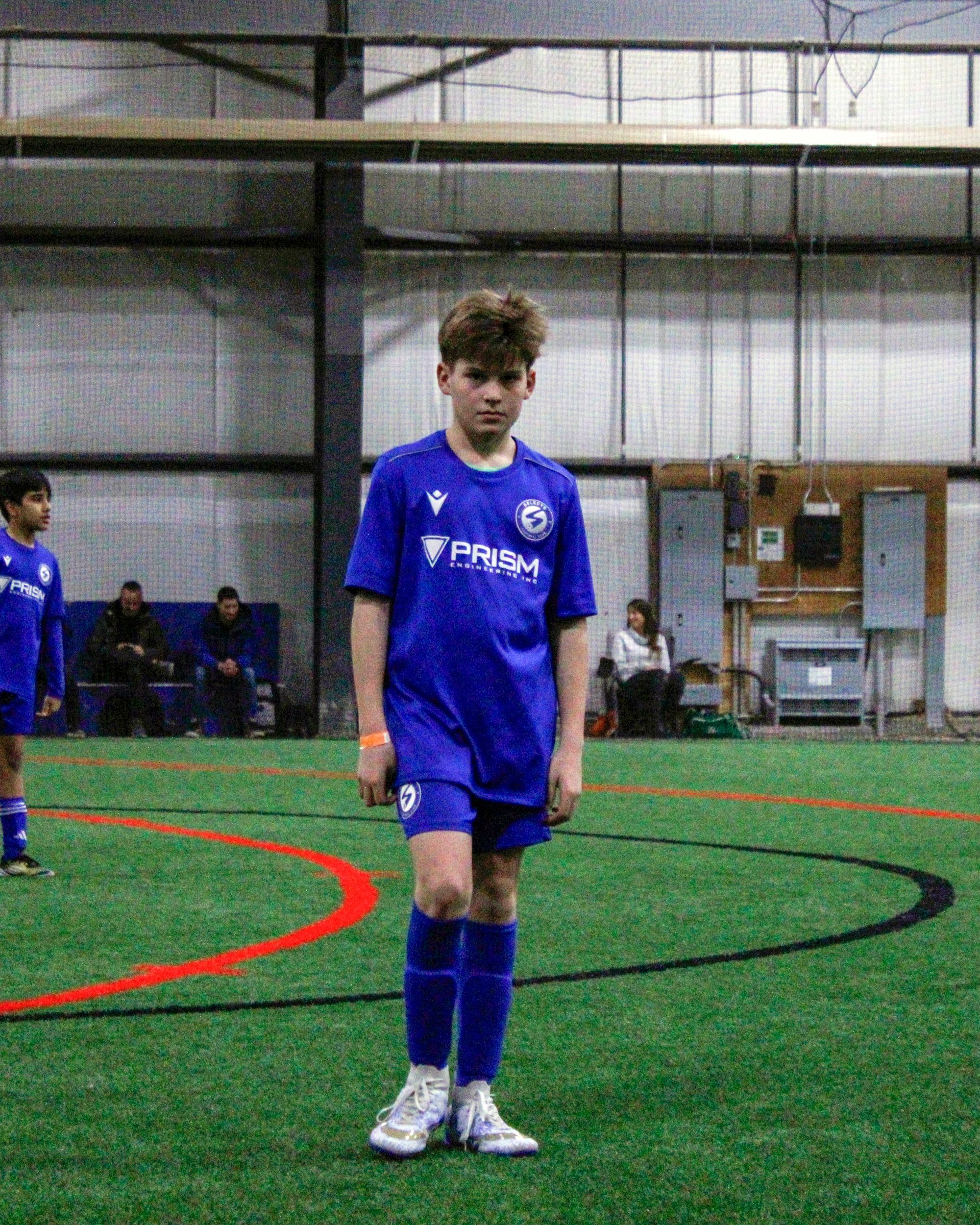 A person in a blue soccer uniform stands on an indoor turf field with a soccer ball at their feet.