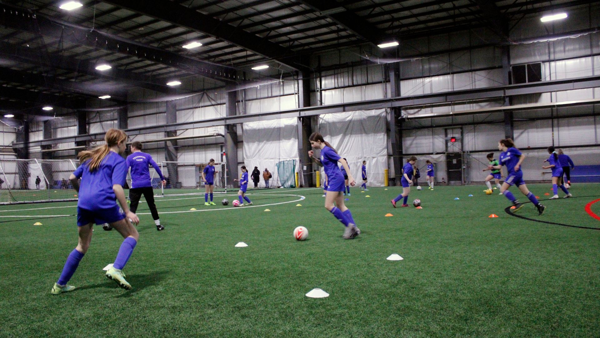 Athletes in blue uniforms practice soccer drills with cones on an indoor turf field.