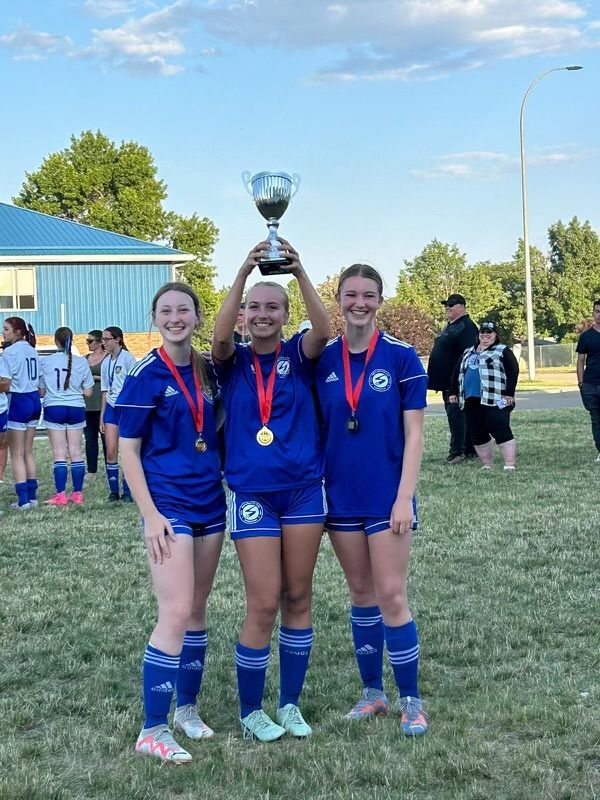 Three soccer players in blue uniforms hold a trophy and wear medals on a grass field, with teammates in the background.