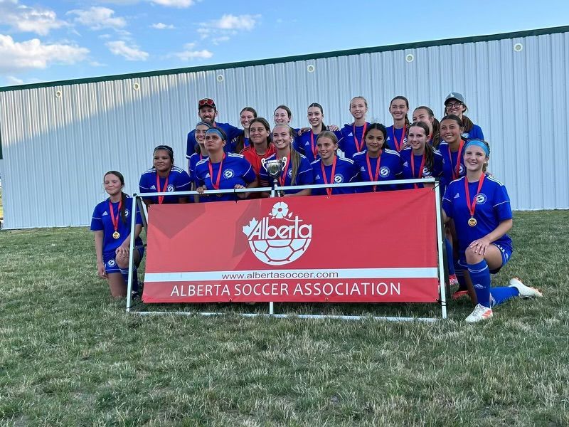 A soccer team in blue uniforms stands behind an Alberta Soccer Association banner, many wearing gold medals, outdoors.