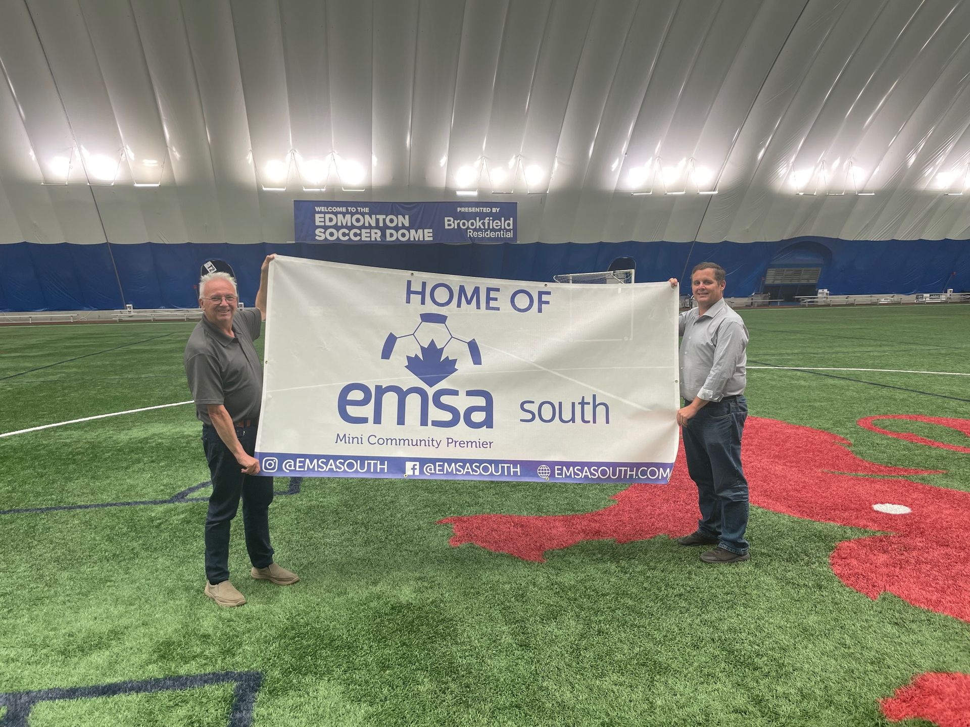 Two people stand on an indoor turf field holding a banner that reads 