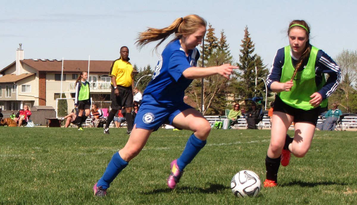 Two soccer players in blue and lime green uniforms compete for the ball on a grassy field with a referee nearby.