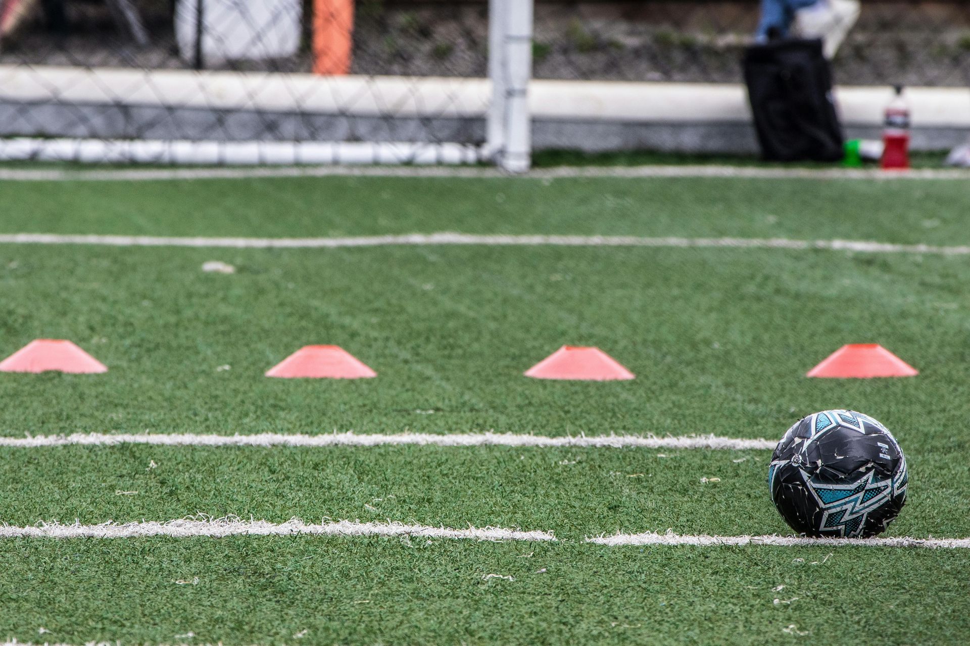 A patterned soccer ball rests on an artificial turf field lined with white stripes and four small orange cones.