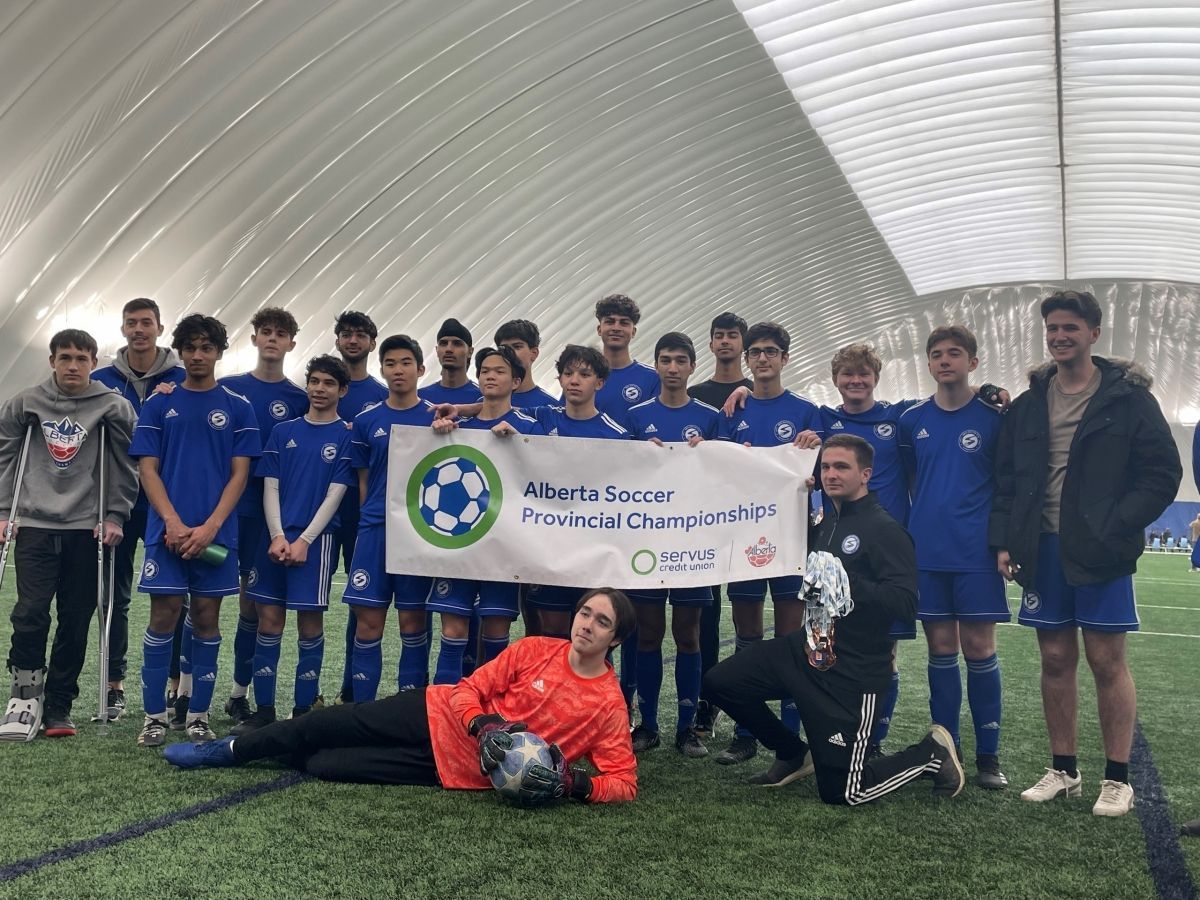 A soccer team in blue uniforms holding a championship banner indoors on a turf field, with one player kneeling in front.