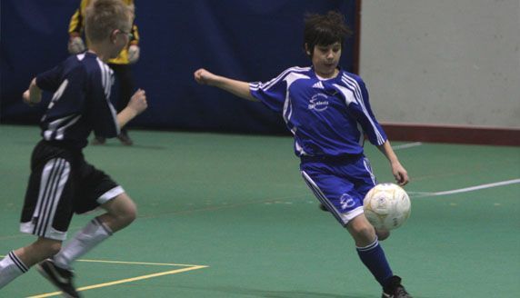 Two youth soccer players in blue uniforms compete for the ball on an indoor court.