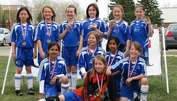 A youth soccer team in blue uniforms holding medals in front of a white banner outdoors.