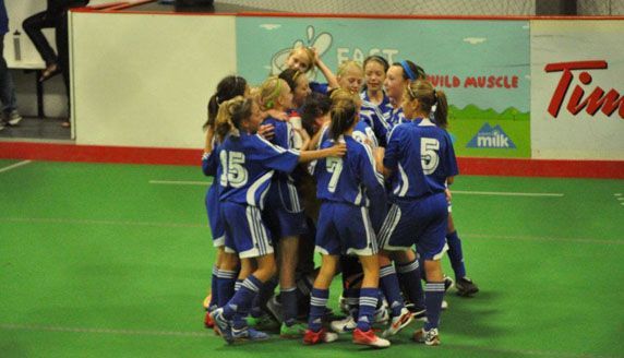 A group of soccer players in blue uniforms huddle together on an indoor field to celebrate a goal.