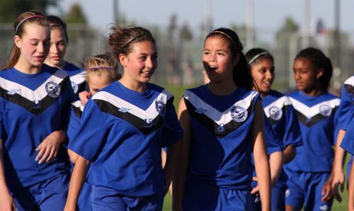 A group of young soccer players in blue and white jerseys walking across a grassy field under a bright sun.