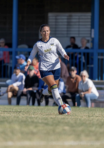 A soccer player in a white jersey and navy shorts dribbling a soccer ball on a grass field in front of spectators.