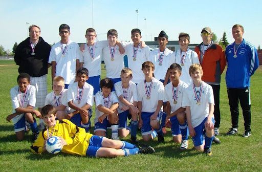 A youth soccer team poses for a photo on a grassy field, wearing white jerseys and blue shorts, with coaches in back.