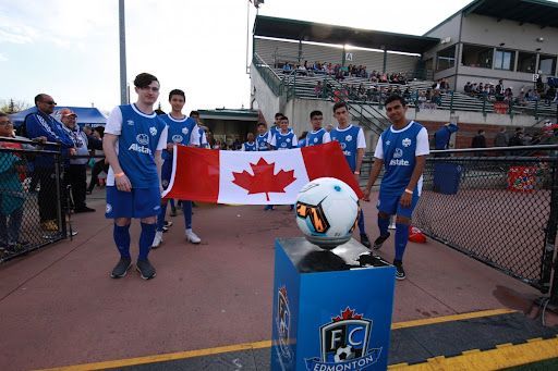 Soccer players holding a Canadian flag near an FC Edmonton stand with a soccer ball on top at an outdoor stadium.