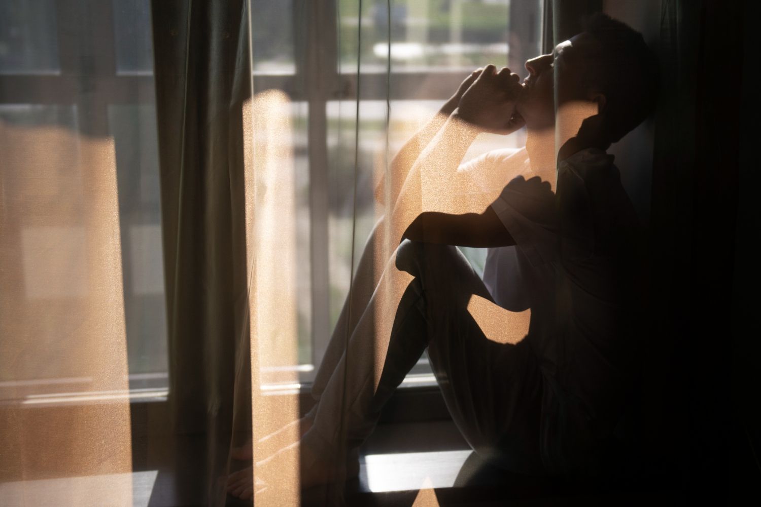 Una persona se sienta encorvada contra una ventana iluminada por el sol, con la cabeza apoyada en la mano. 