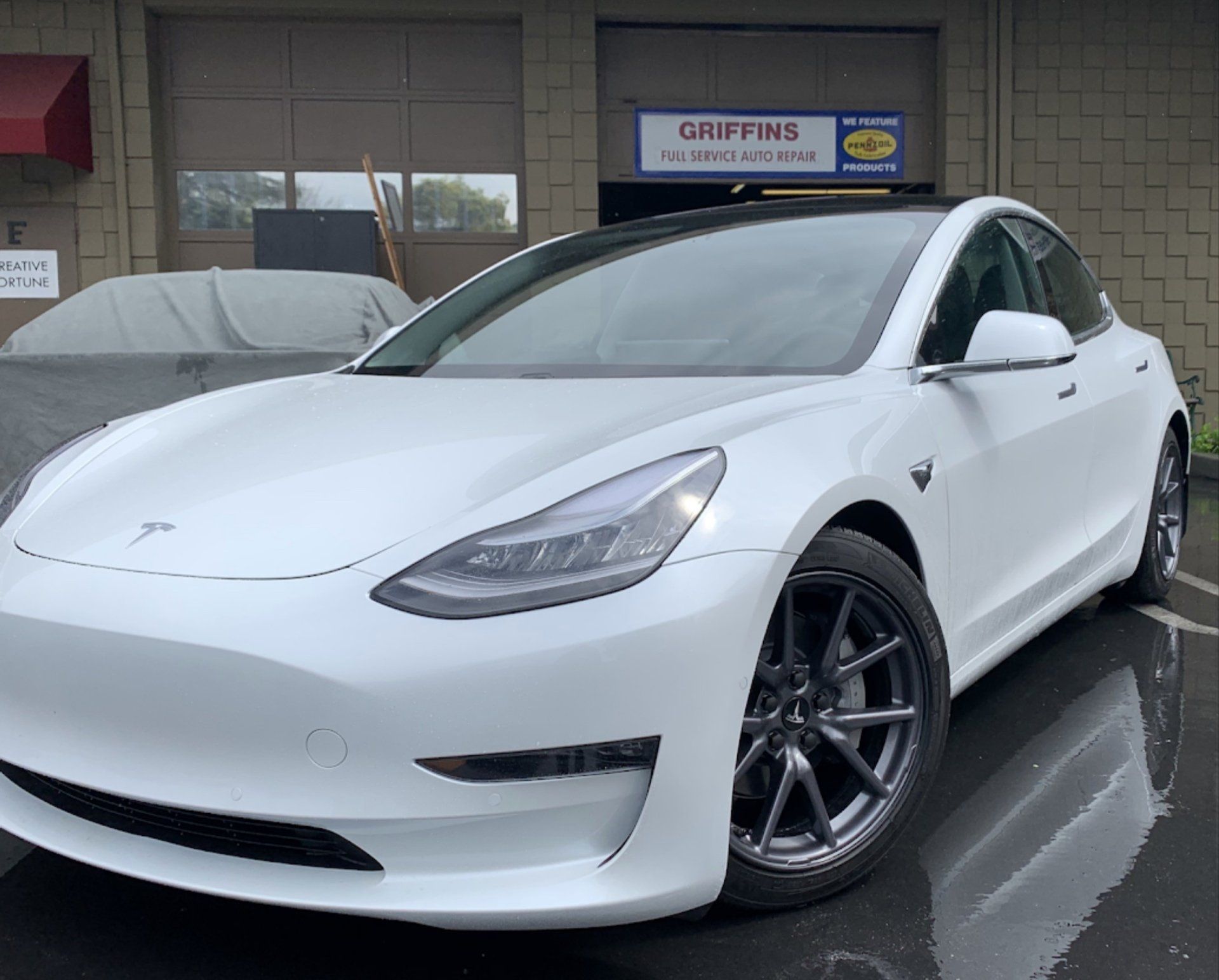 White Tesla sedan parked outside a building on wet pavement, front-left view.