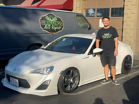Man standing beside a white sports car parked outside a shop with a black van behind it