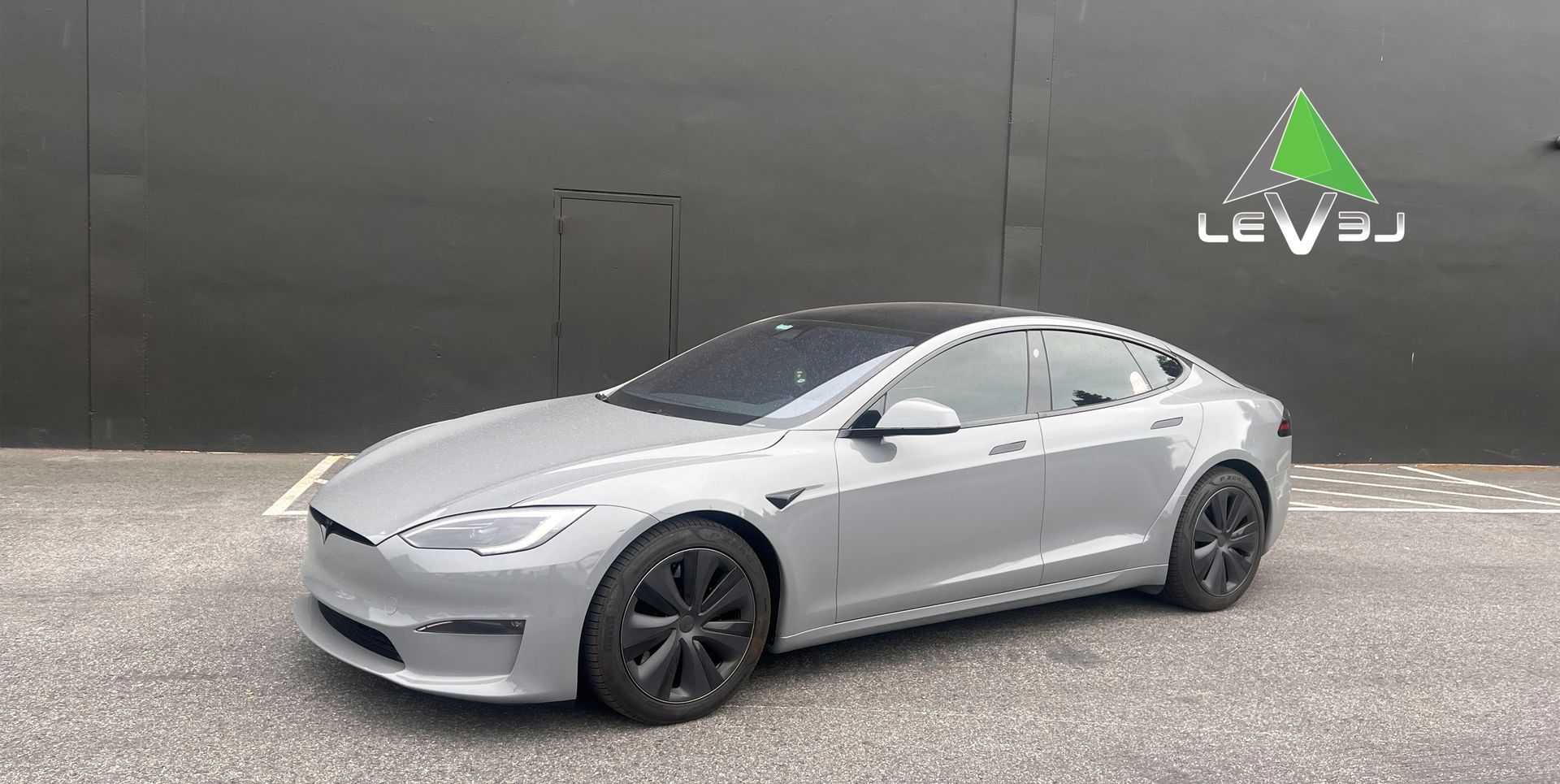 Silver Tesla Model 3 parked on gravel beside a dark wall with a LEVEL logo.